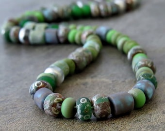 Puede incluir: Un collar de cuentas de vidrio verde, azul y marrón. Las cuentas son de varias formas y tamaños, y algunas tienen una superficie moteada o texturizada.