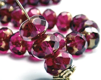 May include: Close-up of faceted, deep magenta beads with gold-colored accents. A needle is threaded through the beads, suggesting jewelry making or beadwork. The beads have a reflective, polished surface and are arranged in a curved line.