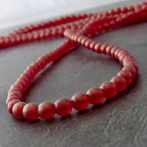 Puede incluir: Un collar de cuentas rojas con acabado mate. Las pequeñas cuentas redondas están ensartadas para formar un collar o pulsera. El color es un rojo intenso y apagado, y las cuentas tienen una superficie ligeramente texturizada.