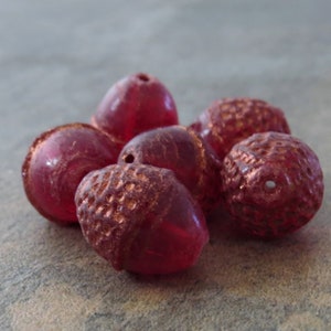May include: A group of six red glass acorn beads with a copper metallic finish. The beads are smooth and shiny, with a slightly textured surface.