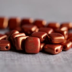May include: A pile of small, square, copper-colored beads with a metallic sheen. The beads have two holes in each corner.