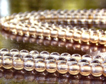 May include: Close-up of two strands of clear, round beads. The beads have a slight amber hue and reflect light, creating a shimmering effect. The beads are strung together, suggesting a necklace or bracelet. The background is blurred, emphasising the jewellery.