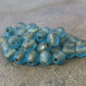 May include: A pile of light blue, frosted glass beads with a metallic sheen. Each bead has a hole for stringing. The beads are irregularly shaped and appear to be handmade, suitable for jewelry making or craft projects.