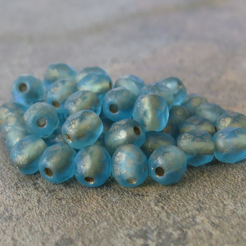 May include: A pile of light blue, frosted glass beads with a metallic sheen. Each bead has a hole for stringing. The beads are irregularly shaped and appear to be handmade, suitable for jewelry making or craft projects.