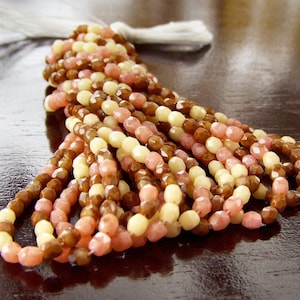 May include: A strand of faceted glass beads in shades of cream, pink, and brown. The beads are arranged in a layered pattern, creating a visually appealing and textured look.