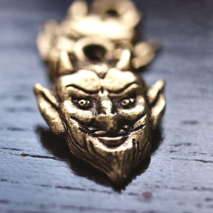 Puede incluir: Un amuleto de metal dorado en forma de cara de diablo con cuernos, barba puntiaguda y una sonrisa traviesa.
