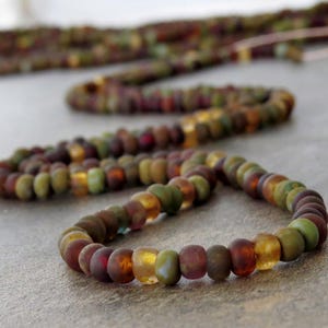 May include: A necklace with a strand of colourful, matte-finish beads. The beads are a mix of earthy tones, including green, brown, amber, and red. The necklace is displayed on a neutral surface, with a blurred strand in the background.