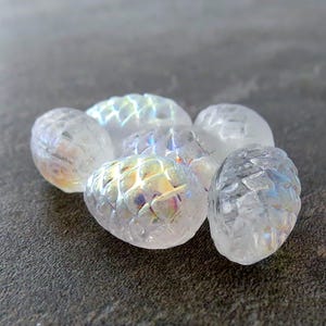 May include: A group of six iridescent white glass pine cone shaped beads. The beads have a textured surface and are arranged in a cluster.
