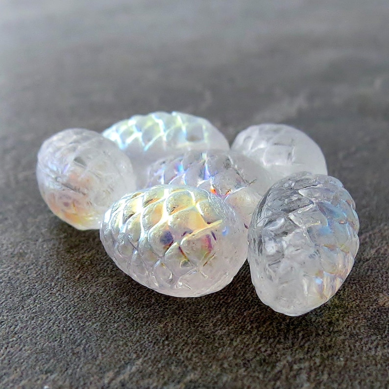 May include: A group of six iridescent white glass pine cone shaped beads. The beads have a textured surface and are arranged in a cluster.