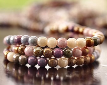 May include: Three beaded bracelets in shades of gray, pink, gold, and purple. The bracelets are made of small, round beads and are stacked on top of each other. The beads have a glossy finish and reflect light.