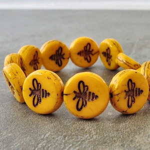 May include: A bracelet featuring round, yellow beads with a distressed finish. Each bead has a dark brown bee design. The bracelet is displayed on a gray surface.