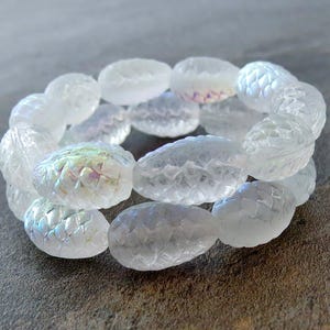 May include: A white iridescent glass bracelet with oval-shaped beads. Each bead has a textured design resembling a pine cone.