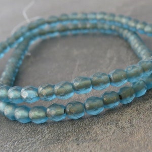 May include: Close-up of a strand of light blue faceted beads. The beads have a slightly iridescent finish and are strung together, creating a delicate and shimmering effect. The beads are round with multiple facets.