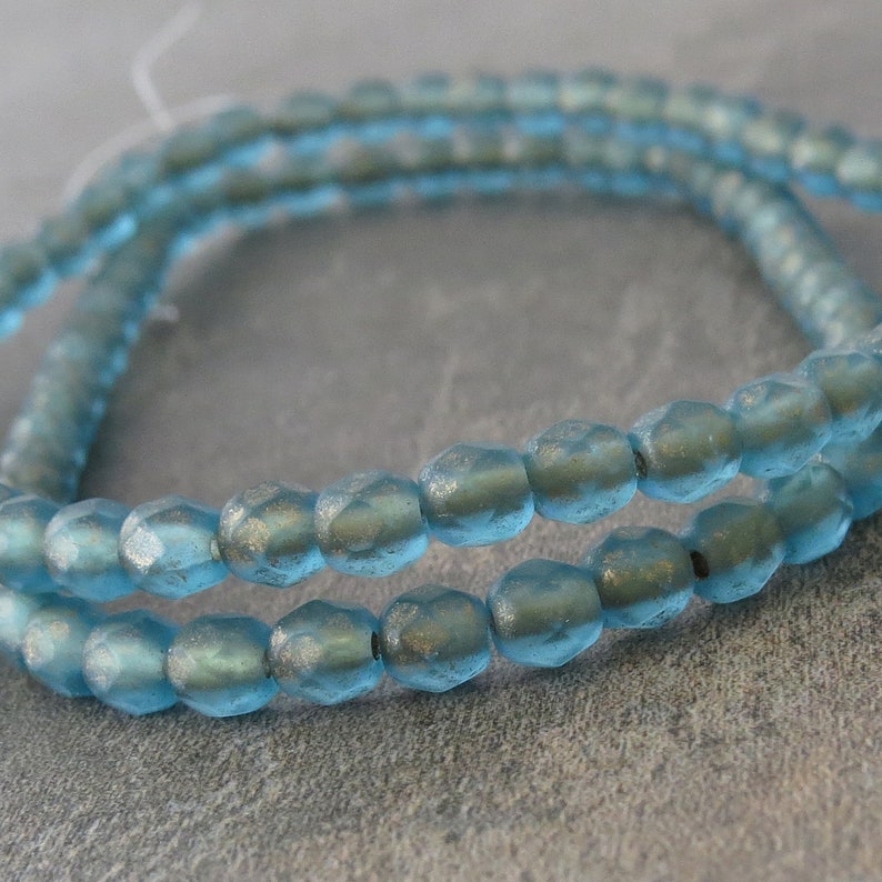 May include: Close-up of a strand of light blue faceted beads. The beads have a slightly iridescent finish and are strung together, creating a delicate and shimmering effect. The beads are round with multiple facets.