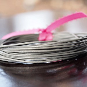 May include: A coil of grey wire, secured with a bright pink ribbon. The wire is tightly wound, and the ribbon is in a bow shape. The background is blurred, with a dark surface.