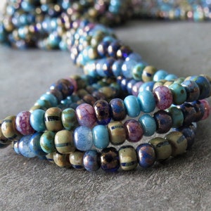 Op de afbeelding: Een close-up van een meerstrengige kralenketting met blauwe, groene en paarse kralen. De kralen zijn klein en hebben verschillende texturen en afwerkingen.