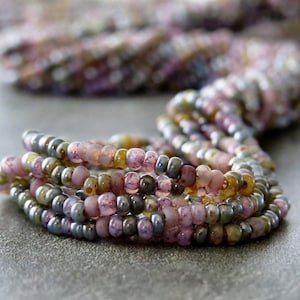 May include: Close-up of several strands of colourful seed beads in shades of pink, purple, green, and yellow. The beads are small and round, with a matte finish. The strands are coiled on a grey surface.