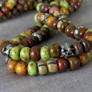 May include: Close-up of strands of artisan beads in various colours, including green, brown, orange, and silver. The beads have a glossy finish and are irregularly shaped. They are strung together, perfect for jewellery making. Each bead is approximately 1 cm in diameter.