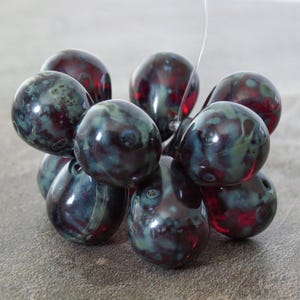 May include: A cluster of dark, glossy beads with a marbled appearance. The beads are primarily dark red and teal, with a reflective surface. The beads are strung together, suggesting they are for jewelry making or crafting.
