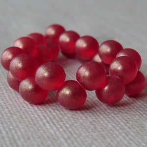 Puede incluir: Un brazalete de cuentas rojas. Las cuentas son redondas y tienen un acabado ligeramente iridiscente.