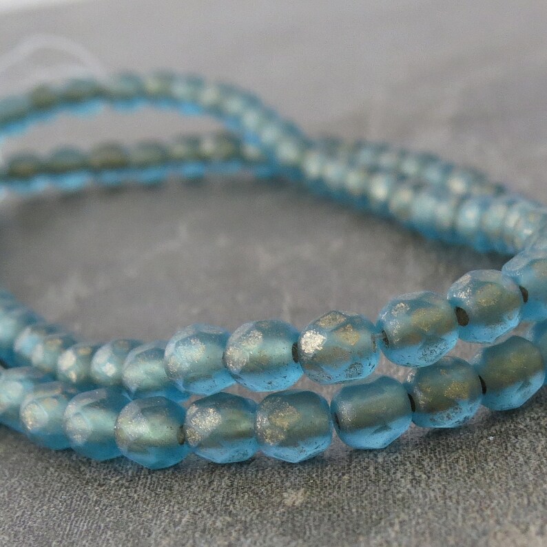 May include: Close-up of strands of light blue faceted glass beads. The beads have a metallic silver finish on some facets, creating a shimmering effect. The beads are strung together, suggesting their use in jewelry making or crafts.