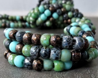 May include: Close-up of three beaded bracelets in shades of turquoise, green, bronze, and gray. The bracelets are stacked, with the beads varying in size and texture. The background is blurred, emphasizing the jewelry.