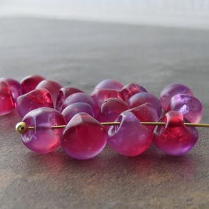 May include: A collection of translucent, irregular, rounded beads in shades of vibrant magenta and deep pink. The beads are strung together on a thin, metallic gold-colored wire that features a small, spherical gold-toned bead at the end.