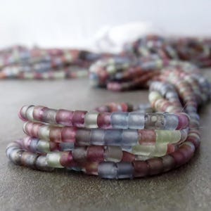 May include: Close-up of several strands of colourful glass beads. The beads are a mix of muted colours, including pink, purple, blue, and green. The beads are strung together, forming a coiled shape. The background is blurred, with more beads visible.