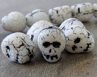 May include: A group of white skull beads with black painted details. The beads are made of a textured material that resembles bone.