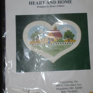 May include: A cross-stitch kit titled "HEART AND HOME" designed by Diane Arthurs. The kit features a heart-shaped design with a house, trees, and the name "drews" and the year "1991". The kit is packaged in a clear plastic wrap.