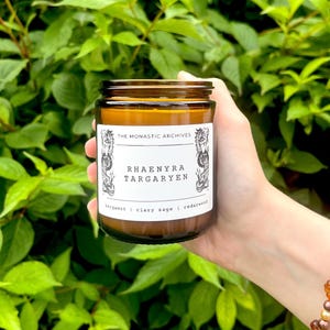May include: A brown glass candle jar with a white label that reads "The Monastic Archives" and "Rhaenyra Targaryen". The label also lists the scents: bergamot, clary sage, and cedarwood.