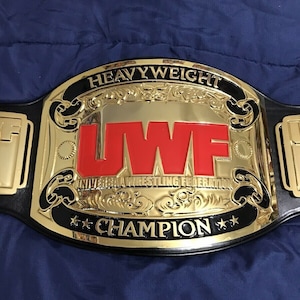May include: A gold and black heavyweight championship belt with the letters "UWF" in red. The belt reads "HEAVYWEIGHT CHAMPION" and "UNIVERSAL WRESTLING FEDERATION". The belt is on a blue surface.