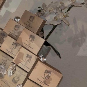 May include: A collection of beige boxes with the text "Growing & Glowing Beauty Bar" and a line drawing of a woman's face. The boxes are arranged on a reflective surface, with clear crystal-like decorations and white orchid flowers.