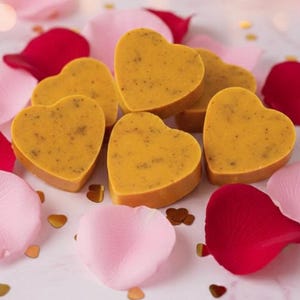 May include: Heart-shaped, golden-yellow soaps with a speckled texture are arranged among pink and red rose petals and gold confetti. The soaps are the focal point, suggesting a theme of self-care or a romantic gift.