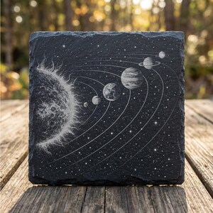 Pu&ograve; includere: Un sottobicchiere quadrato in ardesia nera con una dettagliata illustrazione bianca del sistema solare. Il disegno include il sole, i pianeti e le loro orbite, su uno sfondo stellato. Il sottobicchiere &egrave; esposto su una superficie di legno.