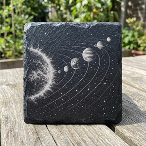 Pu&ograve; includere: Un sottobicchiere quadrato in ardesia nera con una dettagliata illustrazione bianca del sistema solare. Il design include il sole, i pianeti e le orbite su uno sfondo stellato. Il sottobicchiere &egrave; posto su una superficie di legno.