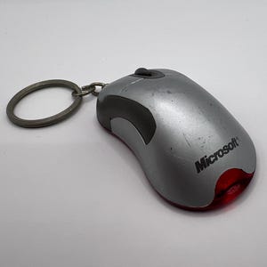 May include: Silver computer mouse keychain with the Microsoft logo. The mouse has a black accent and a red light at the bottom. The keychain ring is silver.