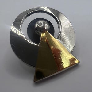 May include: A silver-toned brooch with a gold triangle and a silver circle design. The brooch has a small silver ball at the center of the circle. The brooch is on a white background.