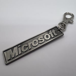 Puede incluir: Un llavero rectangular plateado de Microsoft con el nombre de la marca en relieve en una fuente sans-serif en negrita. El llavero tiene un cierre metálico para sujetarlo. El fondo es una superficie blanca lisa.