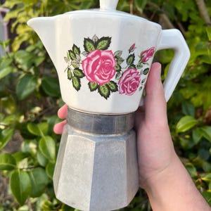 Puede incluir: Una cafetera italiana con una parte superior de cerámica blanca decorada con un patrón de rosas rosas y hojas verdes, combinada con una base inferior de aluminio plateado y una tapa de cerámica blanca a juego con un pequeño asa.