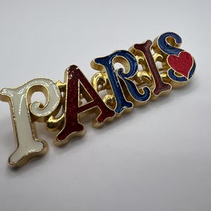 Könnte beinhalten: Eine goldfarbene Metallbrosche mit der Aufschrift "PARIS" in weißer, roter und blauer Emaille. Der Buchstabe "S" zeigt ein rotes Herz. Die Brosche hat ein verziertes Design.