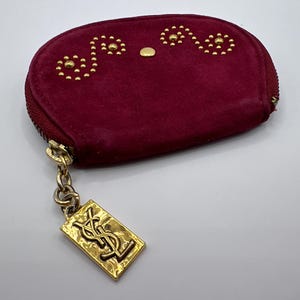 May include: A deep magenta suede coin purse with a gold-tone zip closure. The front features a decorative pattern of small gold studs arranged in two swirling designs. A rectangular gold-tone metal charm with the YSL logo hangs from the zip pull.