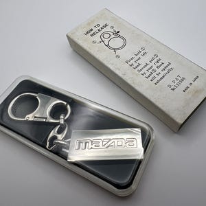 Pode incluir: Um porta-chaves Mazda em metal prateado com um mecanismo de gancho de mola, apresentado numa caixa de plástico transparente. Ao lado encontra-se uma caixa de cartão branca com o texto HOW TO RELEASE, instruções, D. PAT No. 531986 e MADE IN JAPAN.