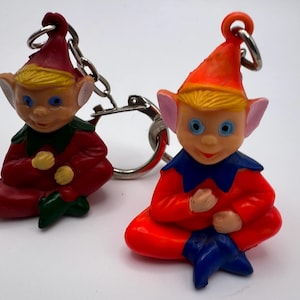 May include: Two elf-shaped keychains with metal rings. One elf wears a red outfit with a dark green collar and hat, while the other wears an orange outfit with a blue collar and hat. Both figures have blonde hair, large ears, and blue eyes.