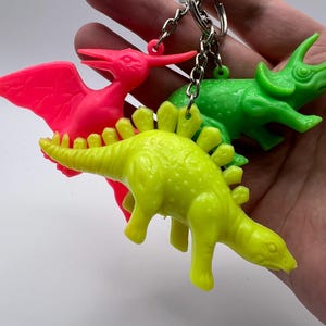 Puede incluir: Un conjunto de tres llaveros de dinosaurios de plástico que incluye un pterodáctilo rosa neón, un triceratops verde brillante y un estegosaurio amarillo lima. Cada figura de dinosaurio está unida a una cadena de metal plateado y una anilla.