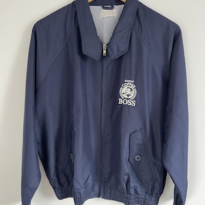 May include: A navy blue zip-up jacket with a collar and two front pockets. The jacket has a white logo on the chest that says "Suntory Coffee Boss". It is hanging on a wooden hanger.