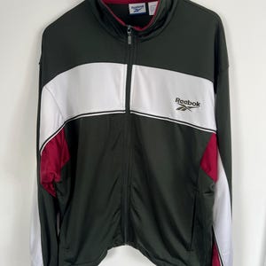 Op de afbeelding: Donkergroen Reebok trainingsjack met een witte streep over de borst en bordeauxrode accenten op de mouwen. Voorzien van een ritssluiting over de hele lengte, een hoge kraag en het Reebok-logo op de borst.