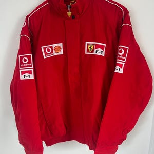 Peut inclure: Une veste de course rouge vif avec un col montant et des manches longues. Le devant et les manches sont ornés d'écussons rectangulaires blancs affichant le logo Vodafone, le logo Shell, l'emblème du cheval cabré Ferrari et le texte F-1.