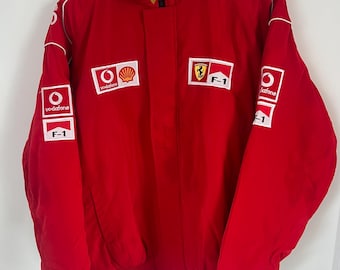 May include: A bright red racing jacket featuring a high collar and long sleeves. The front and sleeves are adorned with white rectangular patches displaying the Vodafone logo, the Shell logo, the Ferrari prancing horse emblem, and F-1 text.
