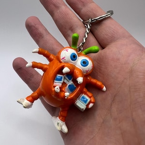 May include: An orange, multi-legged creature keychain with large, wide-eyed features, including blue pupils and red veins. The creature has a wide, open mouth revealing small, square, blue and white teeth-like blocks, and a silver metal chain.
